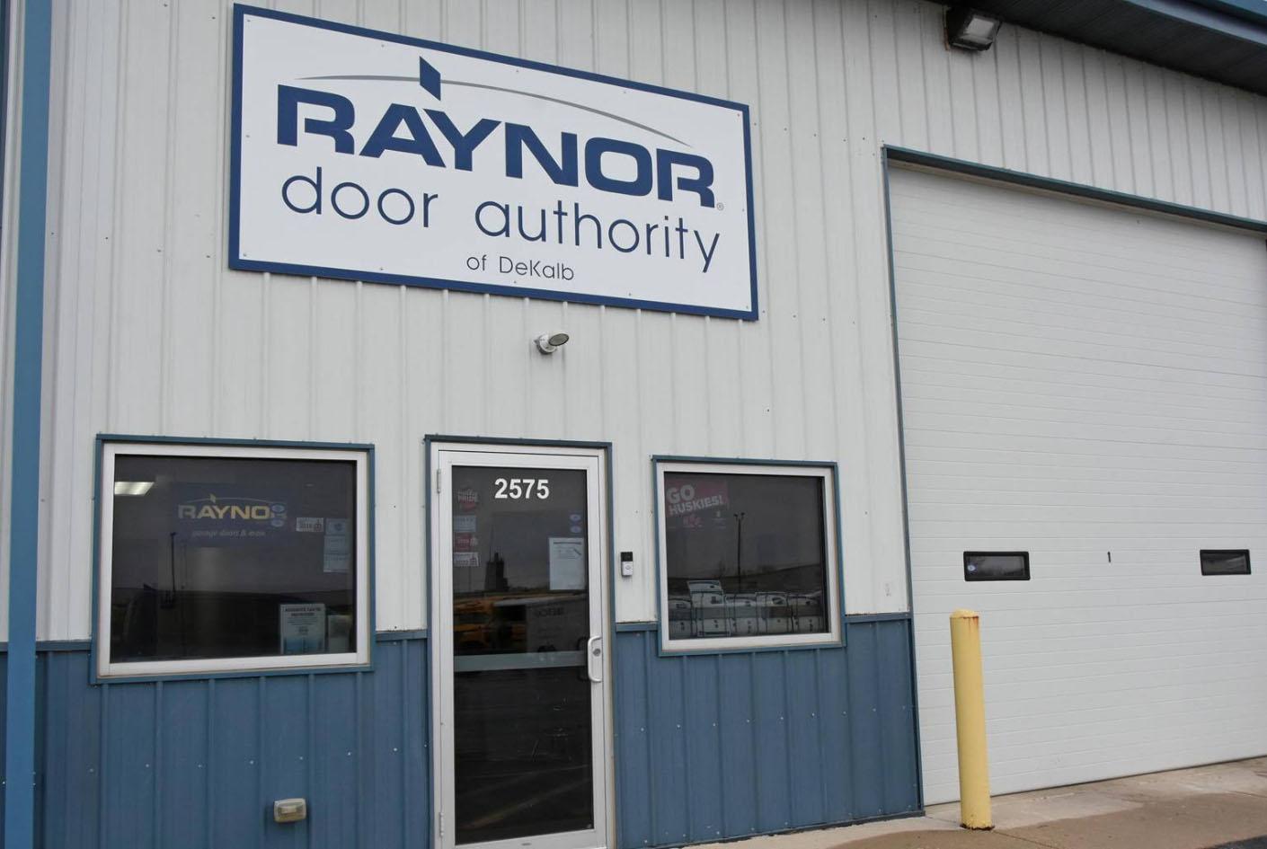 Front view of Raynor Door Authority of DeKalb, featuring a glass door marked 2575, two windows, and a large garage door—your local spot for expert garage door repair in DeKalb. White and blue exterior with yellow bollard.