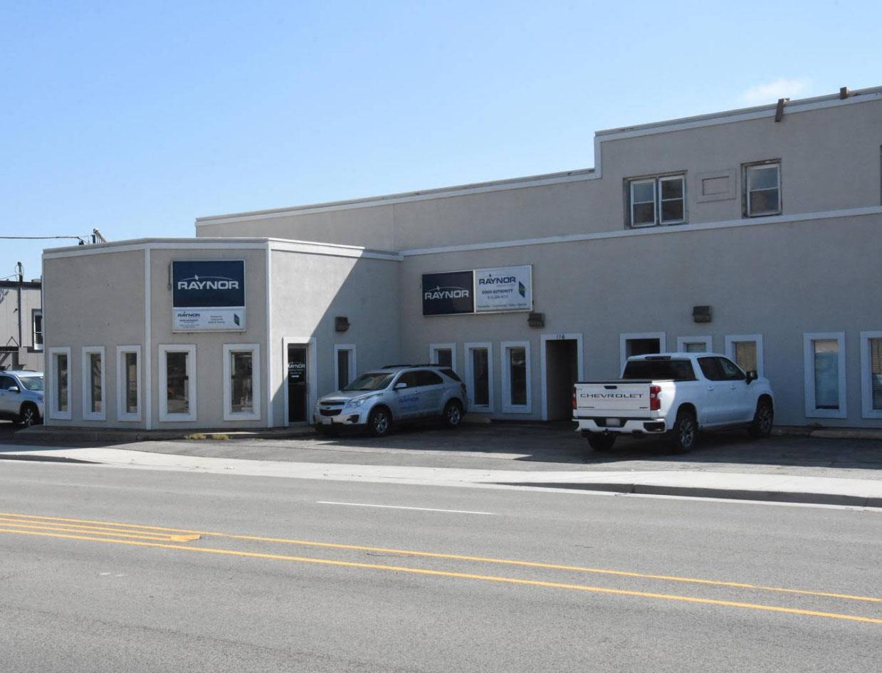 RDA of Sauk Valley A gray commercial building with Raynor signs, several windows, and two trucks parked in front advertises garage door repair Sauk Valley along a sunny street.