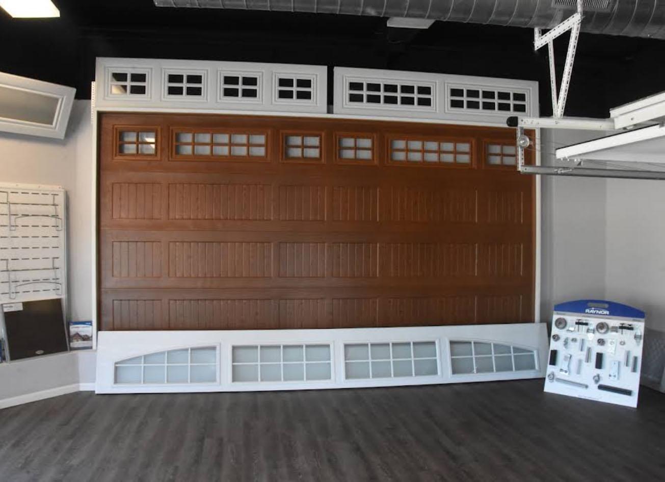 rda sauk valley carriage door Showroom displaying a large brown garage door with windows on top. White door panels, window inserts, hardware samples, and garage door repair Sauk Valley options are arranged along the floor and walls.